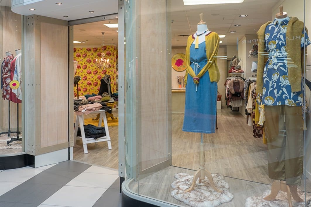 The front shop windows of Kapiti Women's Fashion Store Wallflower. Mannequins standing in the window with Wallflowers bright yellow and pink floral wallpaper at the back of the store